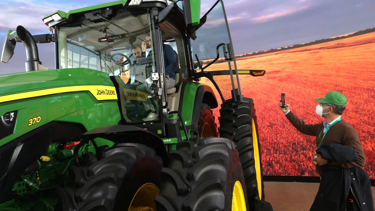 People sit in a tractor in front of a backdrop depicting a farm field. Outside the tractor, a person in a mask takes a picture with his phone.