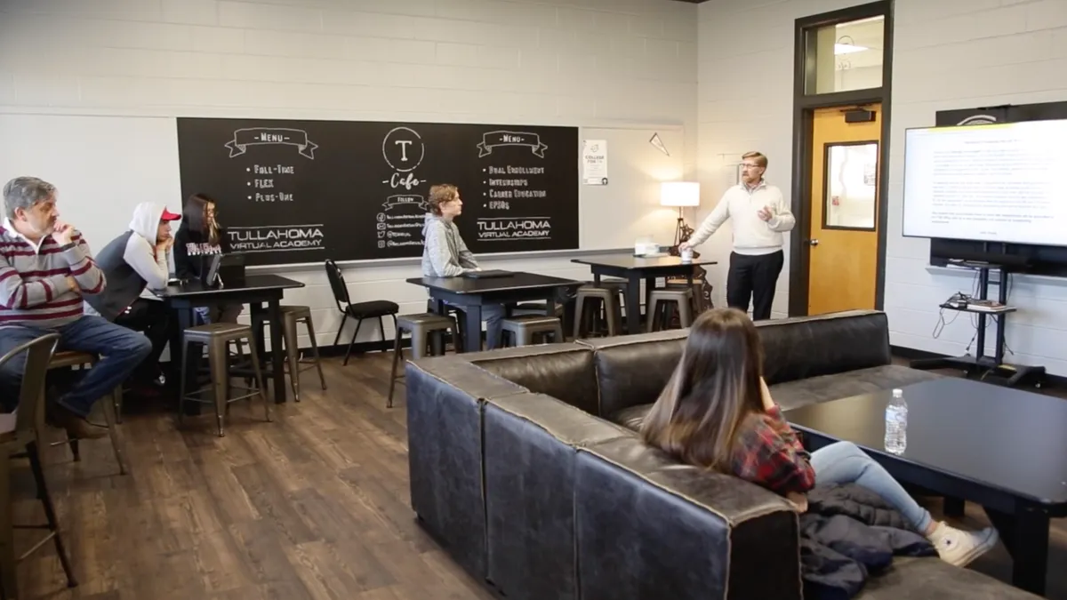 A handful of adults and older students are sitting on a couch and at high-top tables in a room with a blackboard. Another adults is standing near a screen that is projecting words.