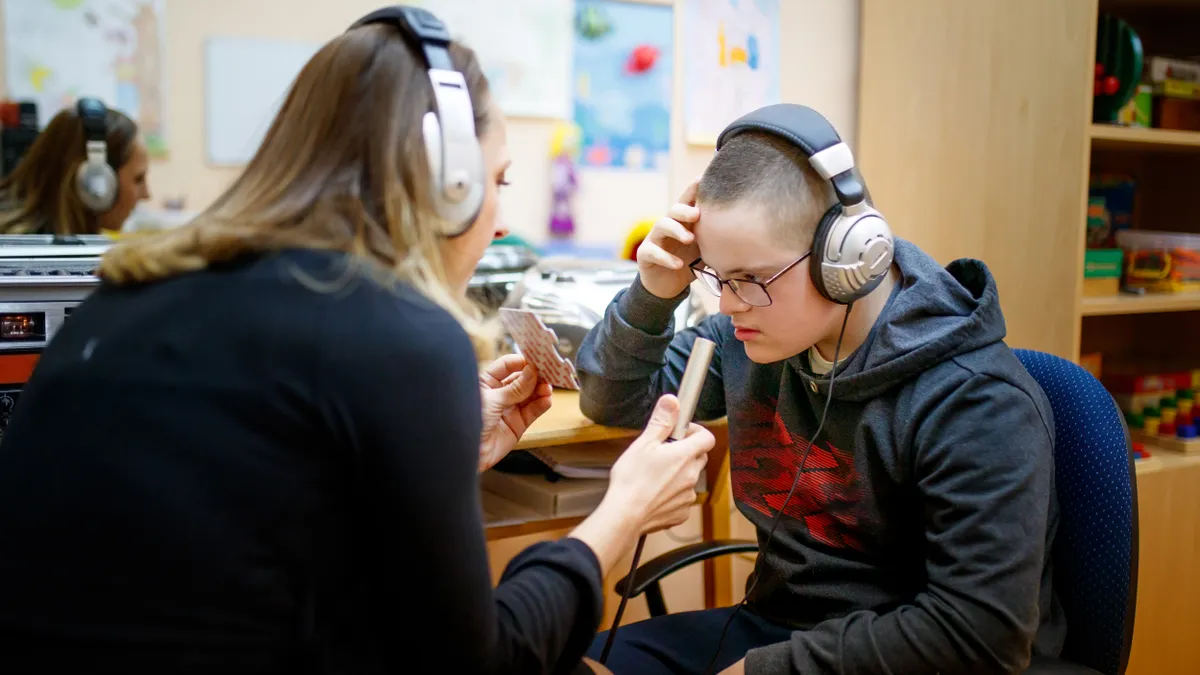 An adult is sitting at a desk with their back toward camera and facing a student who is looking at a flash card in the adult's hands. Both are wearing headphones.