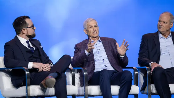 Three men sit on chairs on a stage. the man in the middle is speaking