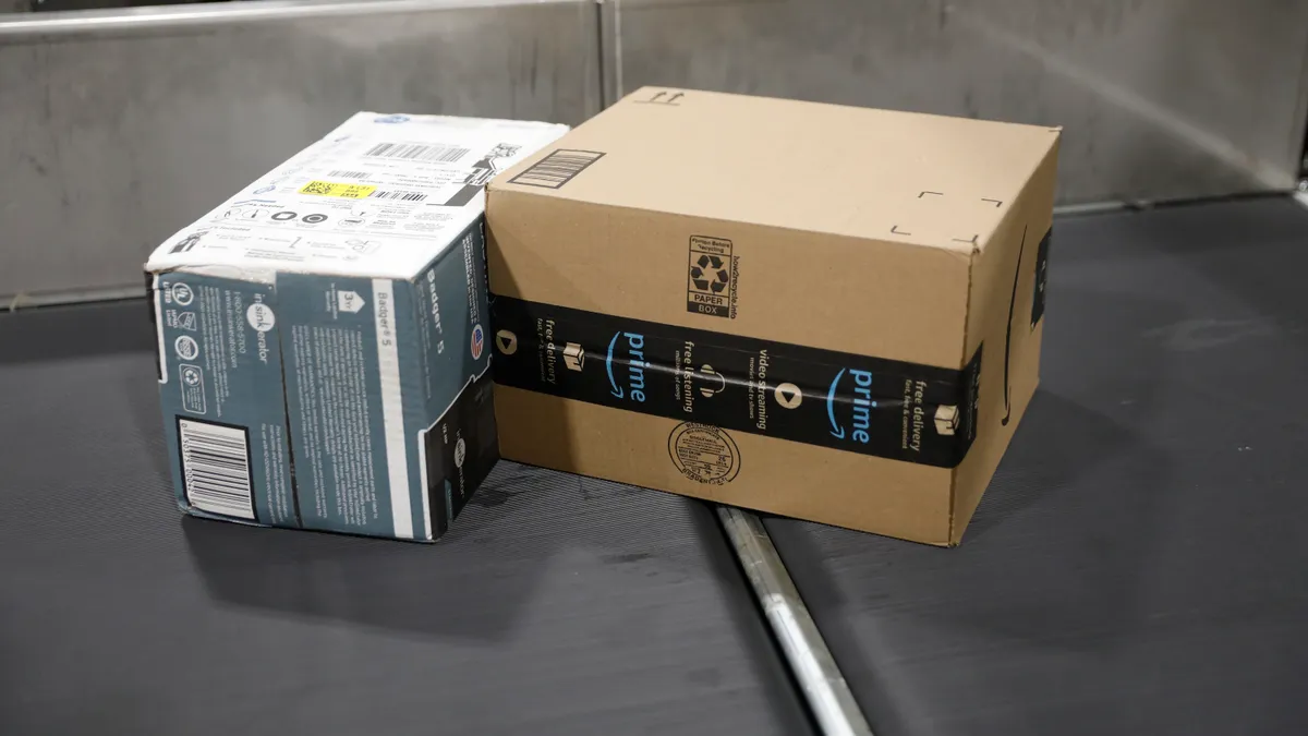 Two boxes on a conveyor belt ready for shipment. The left one is a product box with a shipping label and the right one is an Amazon branded box.