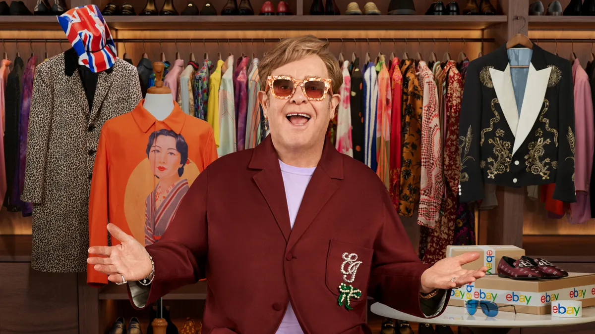 A person in large sunglasses wears a brown jacket and stands smiling in front of a rack of clothing.