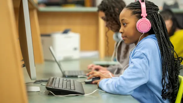 A student wearing pink headphones looks at a desktop computer in a school library computer lab, surrounded by other students working on laptops.