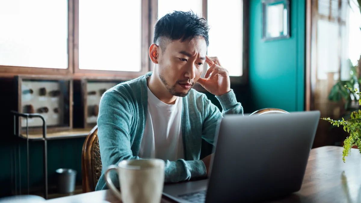 Worried Asian man with his hand on head, using laptop computer at home, looking concerned and stressed out