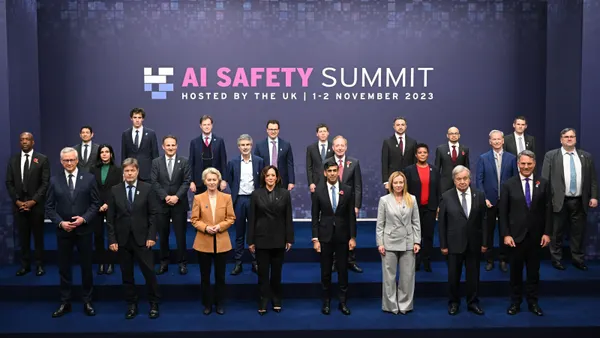More than two dozen world leaders, tech executives, and experts stand in several rows on blue carpeted steps in front of a backdrop that says "AI Safety Summit"