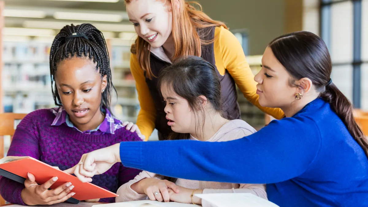 Four students sit at a desk in a school library and all are looking at an open book one student is holding.