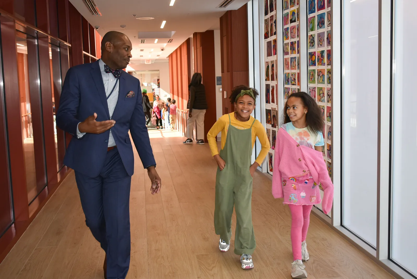 An adult and two students walk through a school hallway. On one side of the hallway student artwork is displayed on the wall.