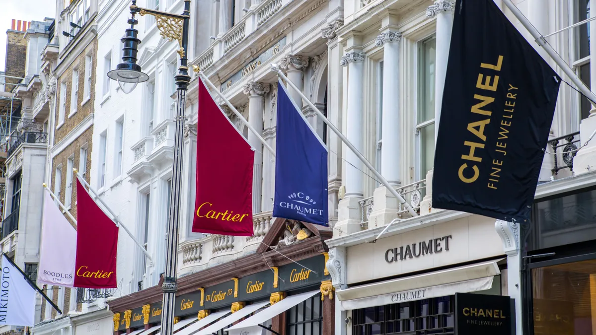 A row of flags branded with luxury names flies on storefronts on a sunny day.