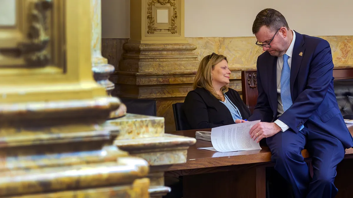 A white man in glasses and a suit sits on a desk while reading through a stack of papers.