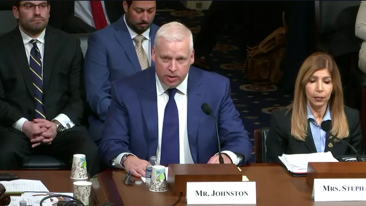 A person in a blue suit and tie, seated at a table, speaking into a microphone.