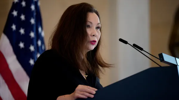 A woman stands at a podium and speaks into a microphone. Behind her is an American flag.