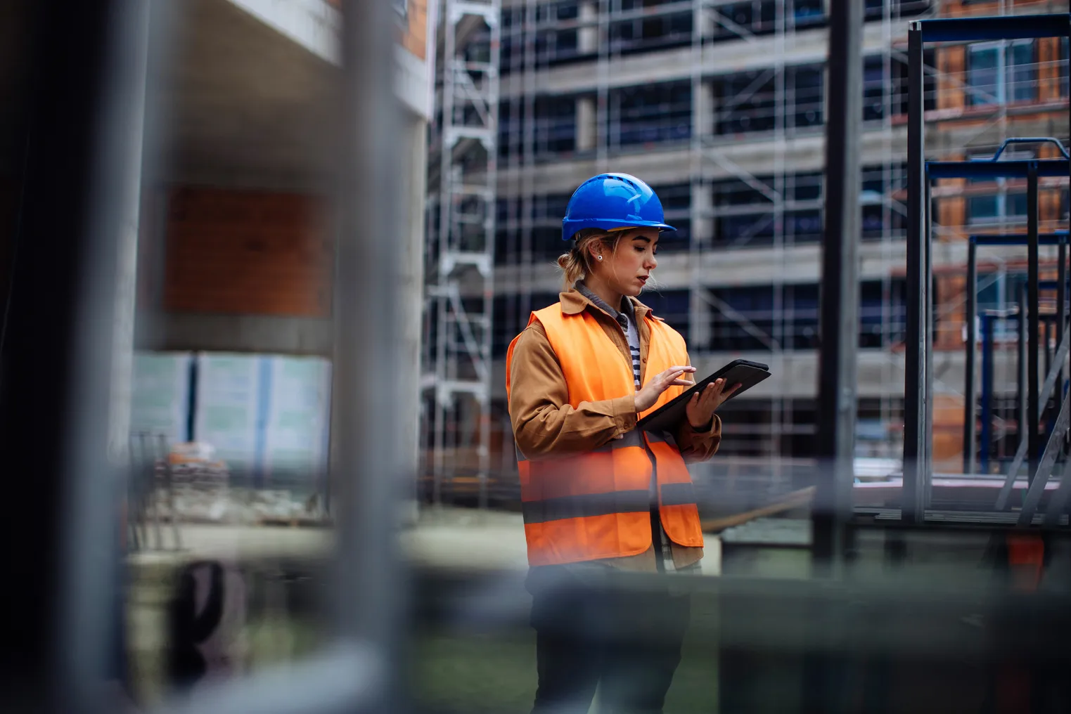 A woman with a safety vest in a hardhat looks at a tablet on a construction site. She is looking at it intently.