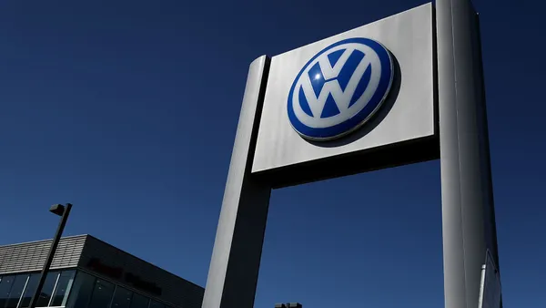 A Volkswagen sign jets out into the blue sky as a dealership building is seen in the lower-left corner of the image.