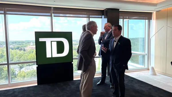 People wearing suits talk near a "TD" sign, in front of a bank of windows.
