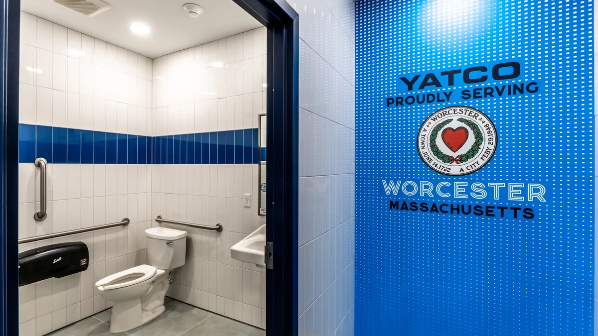 A photo of the interior of a store, showing a hallway and a bathroom through an open door. The bathroom has blue and white tile. A sign on the wall says Yatco. Proudly serving Worcester. Massachusetts.