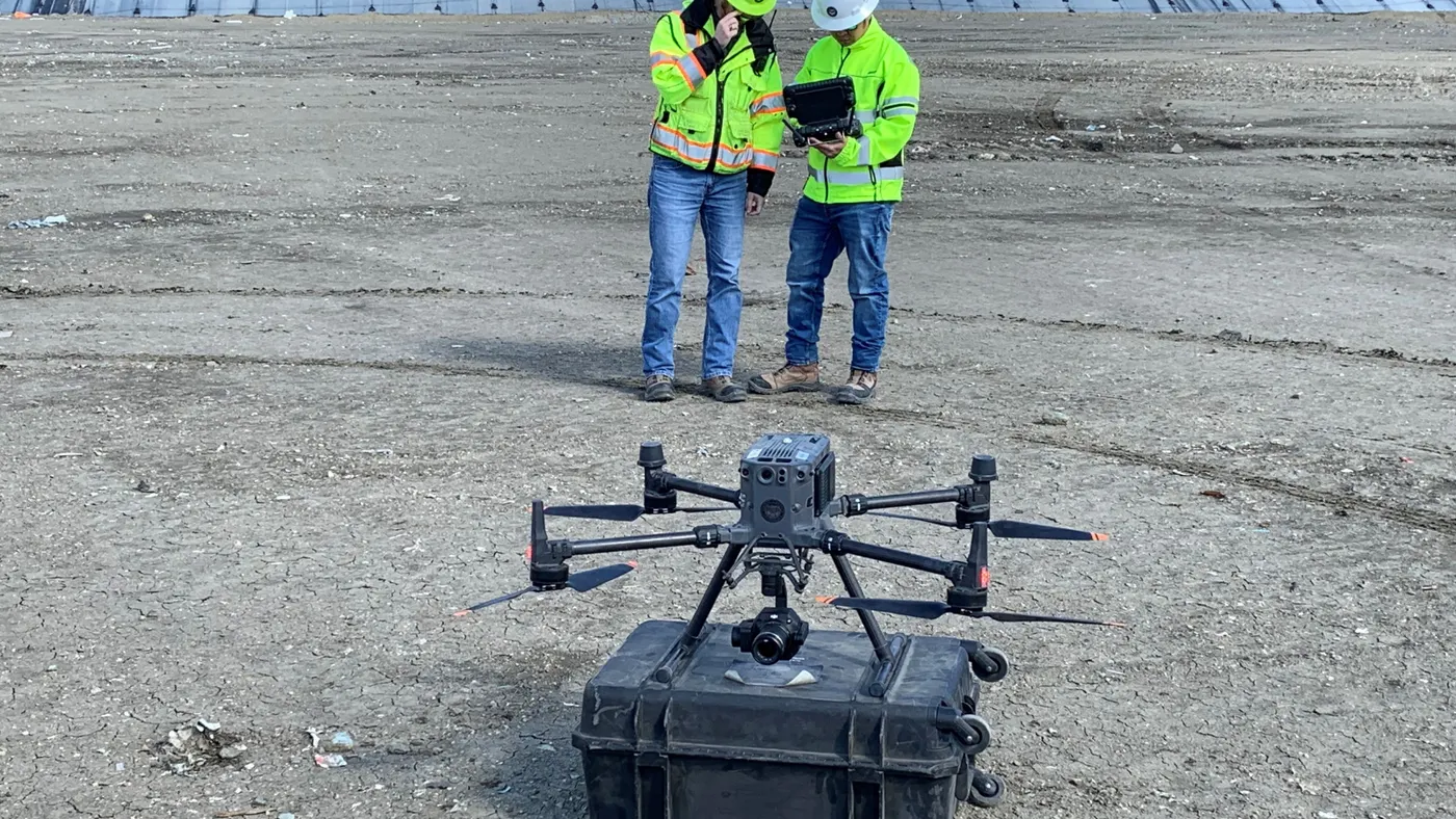 A drone rests atop a carrying case while two people wearing hard hats and construction vests consult a tablet behind it
