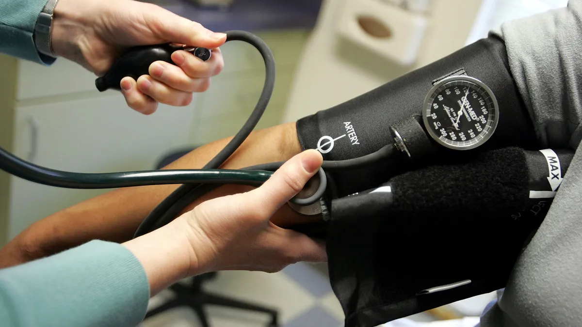 A person holds out their arm with a blood pressure cuff wrapped around it while another person inflates it.