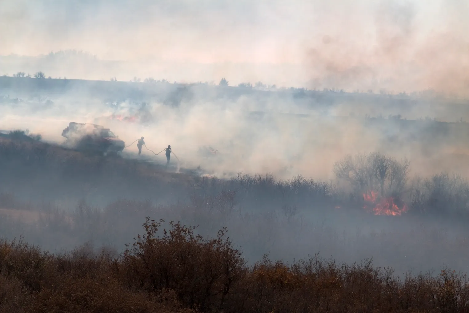 As smoke rises into the air, a pair of firefighters near a vehicle pull a hose and spray water on burning grass and brush.