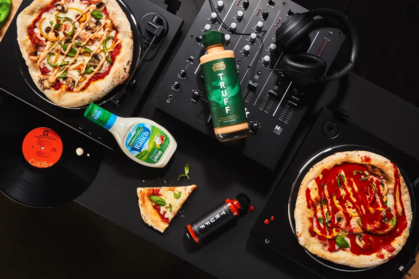 Bottles of Spicy Truffle Ranch, Hidden Valley Ranch and Truff hot sauce lie on an audio mixer. Pizzas sit on two record turntables, and a pair of black headphones and a slice of pizza sit nearby.