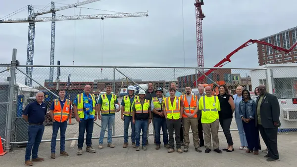 A group of people in construction gear pose for a photograph on a jobsite below cranes.
