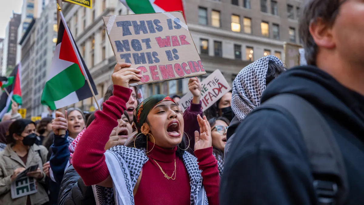 A New York City high school student wearing a keffiyeh holds a sign reading "This is not war this is genocide."