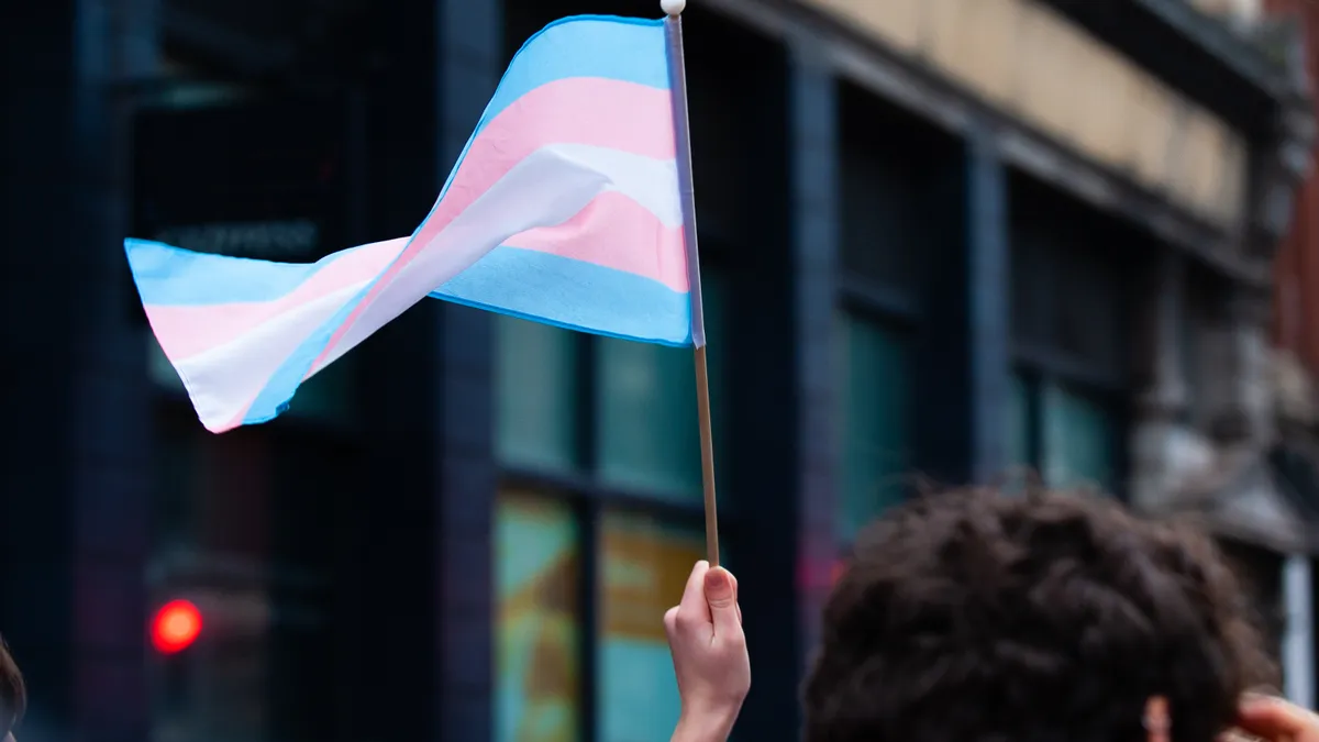 A hand holds up a small transgender pride flag in a Bristol Street. The transgender pride flag is a symbol of pride for the trans community. The blue and pink stripes represent traditional colours for