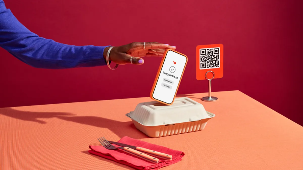 An arm holds a smartphone over a takeout box that is on a red table. On the right is a QSR code.