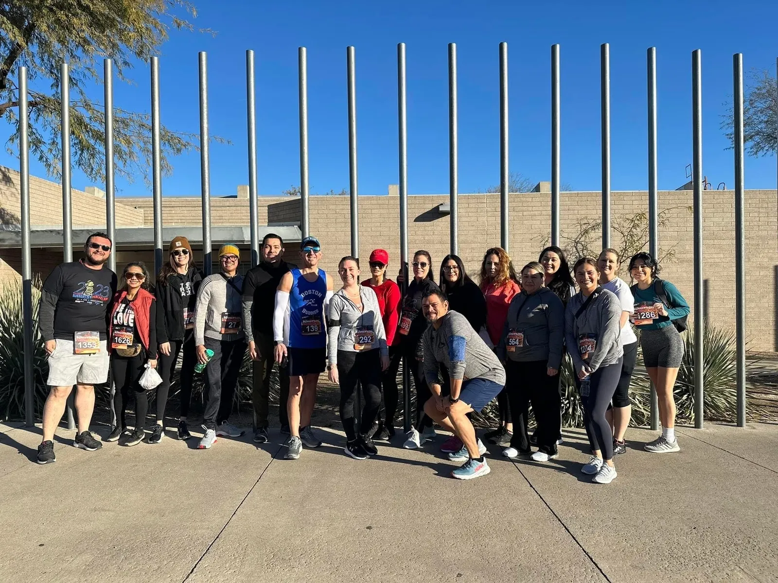 A group of 17 middle school staff members stand in front of a series of metal posts outside of a beige building in Arizona after running a marathon.