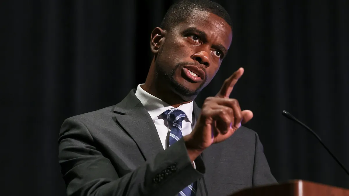 A person stands at a podium and gestures with a raised finger.