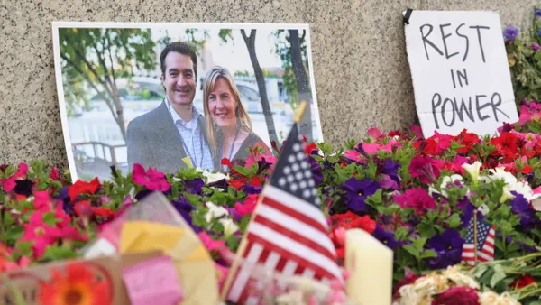 Photo of a couple against a stone wall behind flowers, surrounded by miniature flags and next to cardboard sign that says "REST IN POWER."