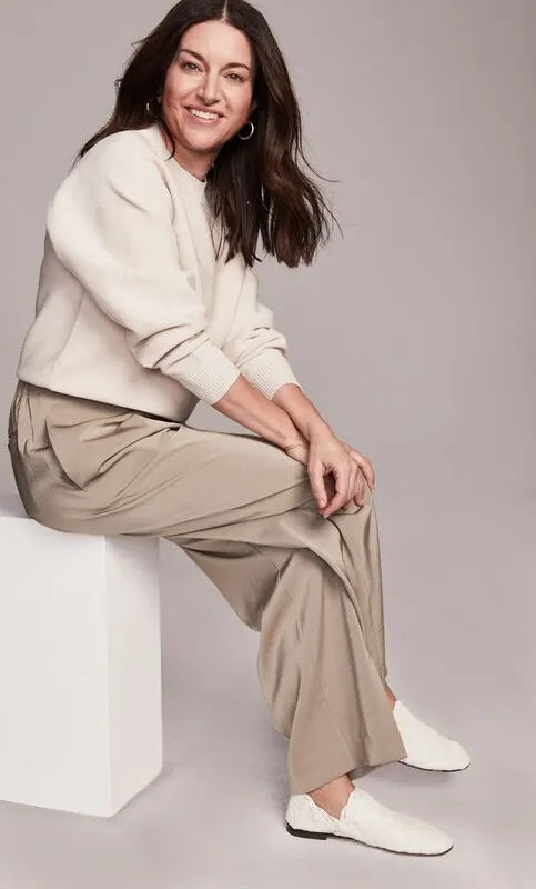 A person in a cream sweater and beige pants sits in front of a light background.