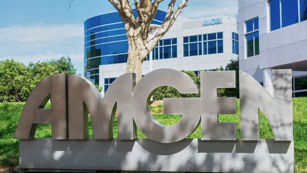 Shrugs and trees surround a large metal Amgen sign displayed outside a modern office building. 