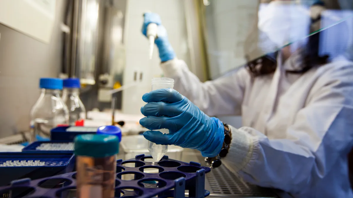 A researcher with blue gloves holds a pipette tool in the right hand and a test tube in the left hand.