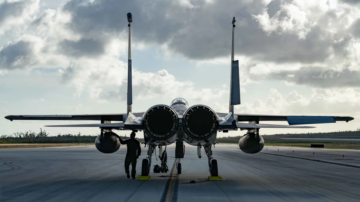 A pilot inspects his fighter jet in the foreground, as the runway frames the jet. There is sky and grass surrounding the runway and jet.