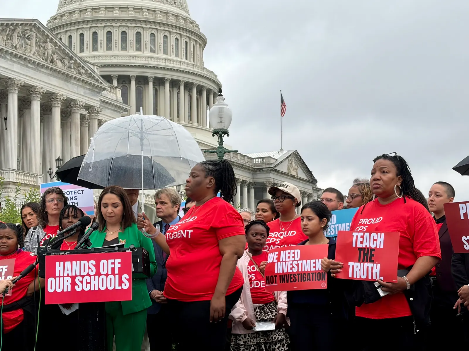 Several people are standing outside the U.S. Capitol. Some are holding umbrellas and signs. One person is speaking at a podium.