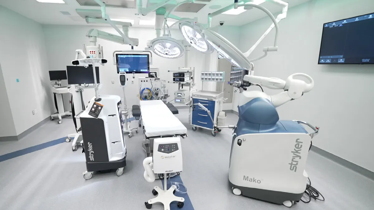 An operating room has a table surrounded by screens and several overhead lights, and a surgical robot that says "Mako"