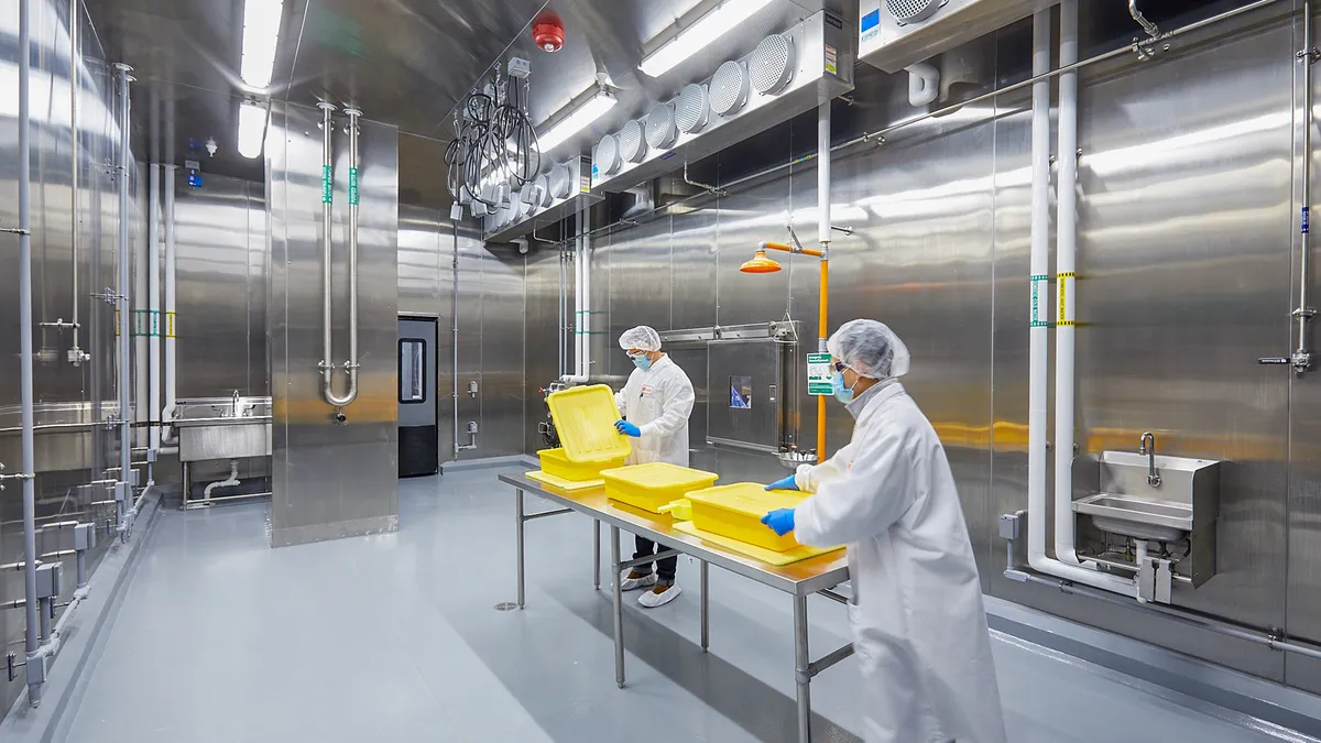 People in food safety protective gear handle yellow plastic tubs on a metal table in the middle of a room with metal walls, tubing and fans.