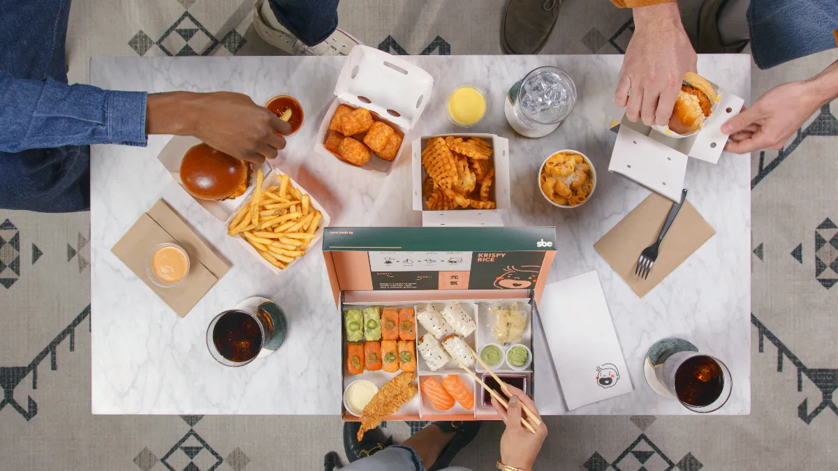 A white table with hands reaching out toward food items like hamburgers, sushi and French fries.