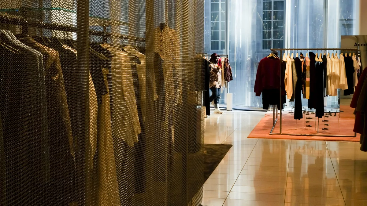 A rack of clothes to the left is partially hidden by a screen. Two other racks of clothes, reflected in the shiny floor, stand in front a huge window.