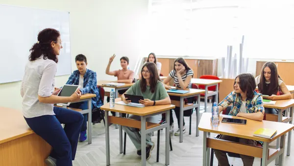 Teenage boys and girls listen to a teacher in a classroom. The teacher is holding a tablet.