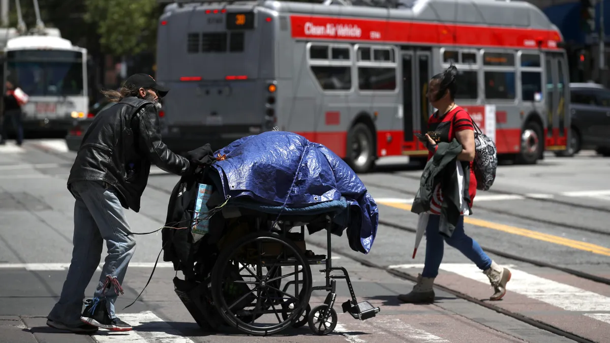 A person in a leather jacket, jeans and baseball cap pushes a cart with a tarp over it while walking across a crosswalk. Another person walks in the opposite direction on the crosswalk. Behind them is a bus and street signs.