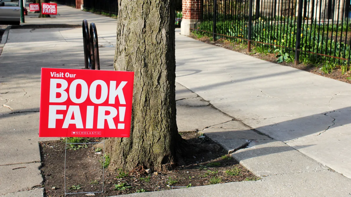 A row of Scholastic book fair signs are planted in the ground next to a sidewalk