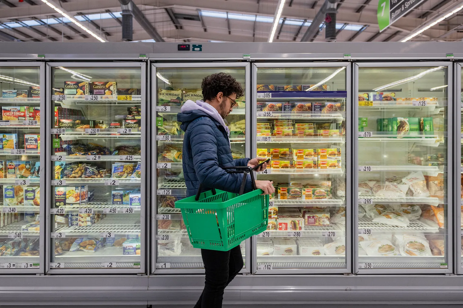 A person holding a green shopping basket checks their phone in the freezer isle.