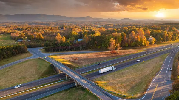 A highway overpass junction with traffic, including a tractor-trailer and passenger vehicles, at sunset.