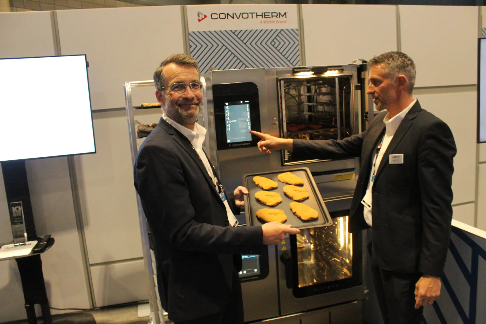 Two men display a tray of schnitzel in front of an oven.