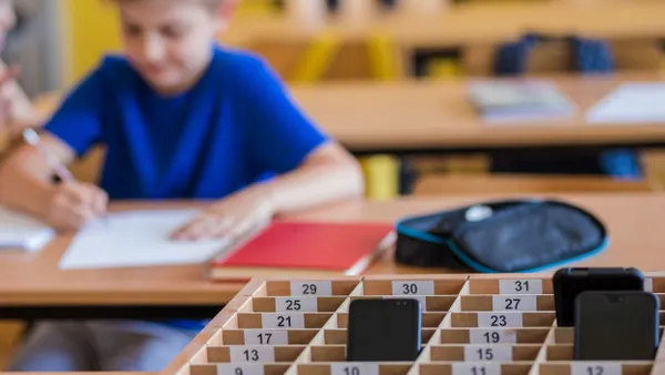 A box of cell phones sits in front of a student completing an assignment at their desk in a classroom.