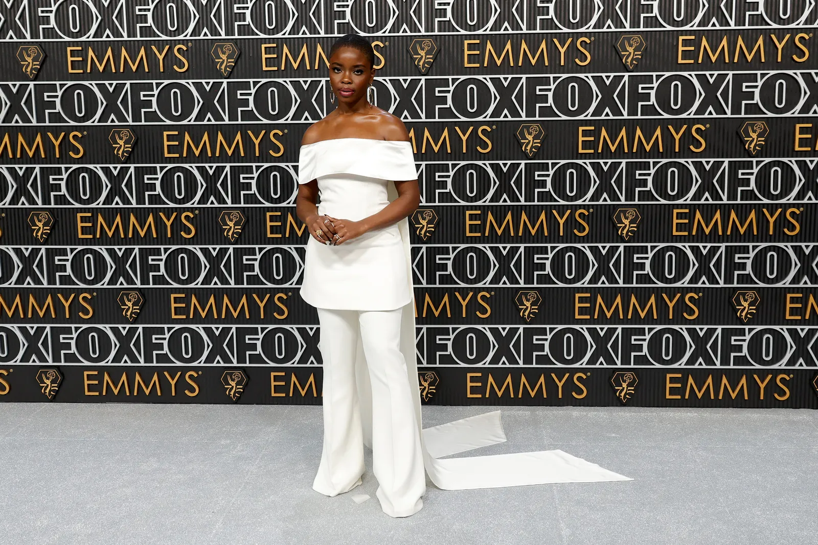 A person in a white pantsuit with a flowing train poses for the camera.