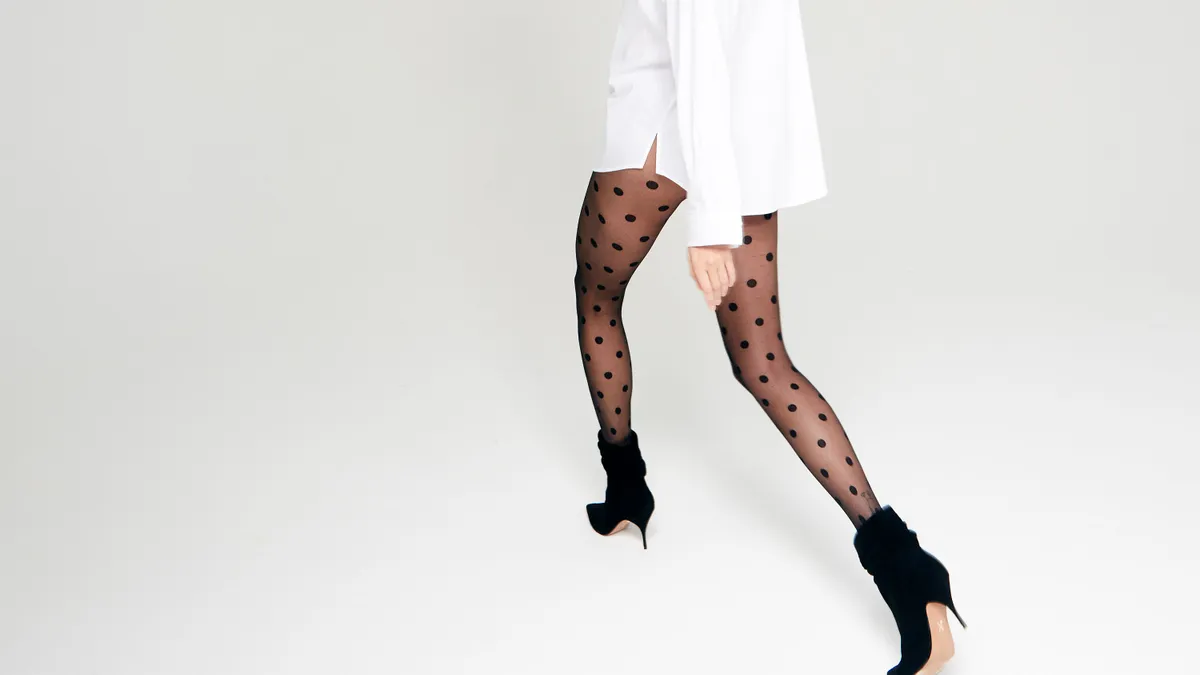 A person in a large white top, polka dot tights, and black Brian Atwood scrunch booties walks away from the camera looking back toward it.