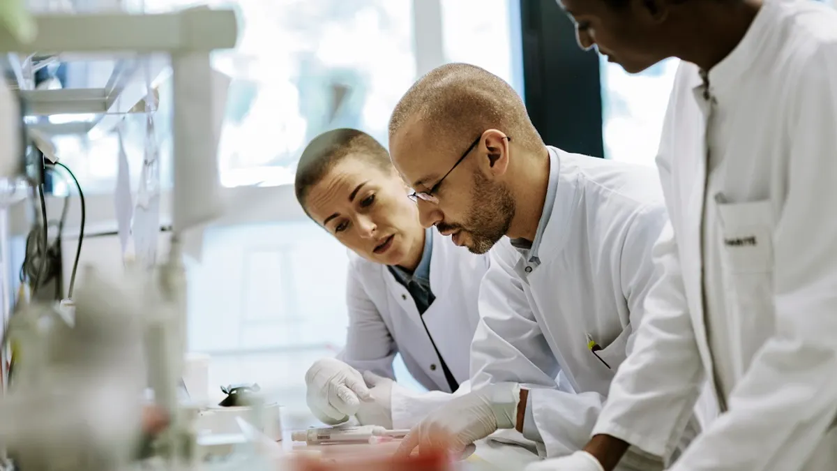 Three scientists in lab coats look at something at a work table.