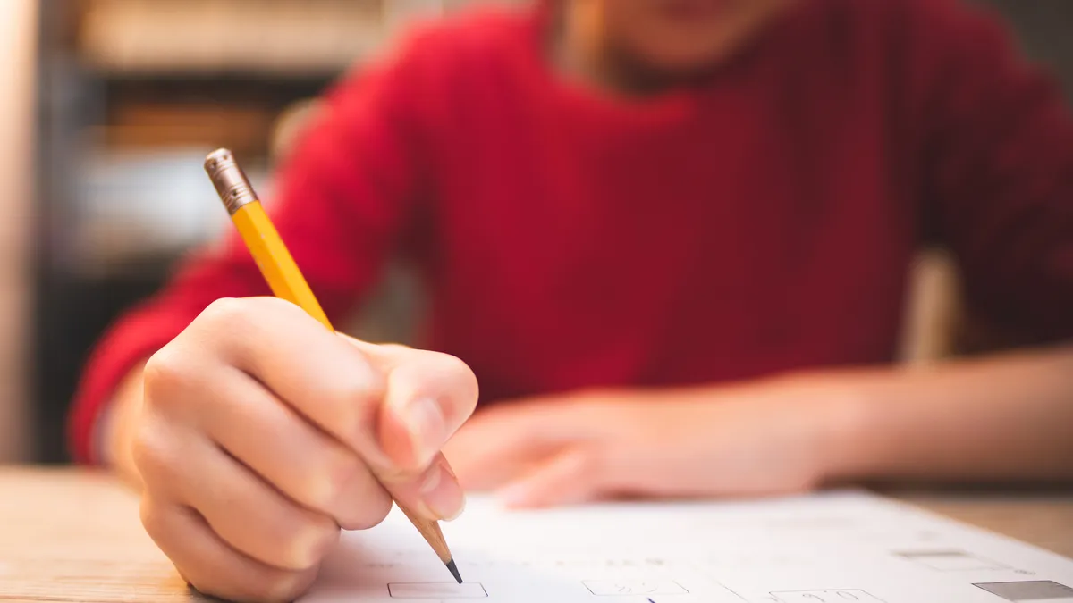 Camera has close-up of pencil being held by person sitting at a desk